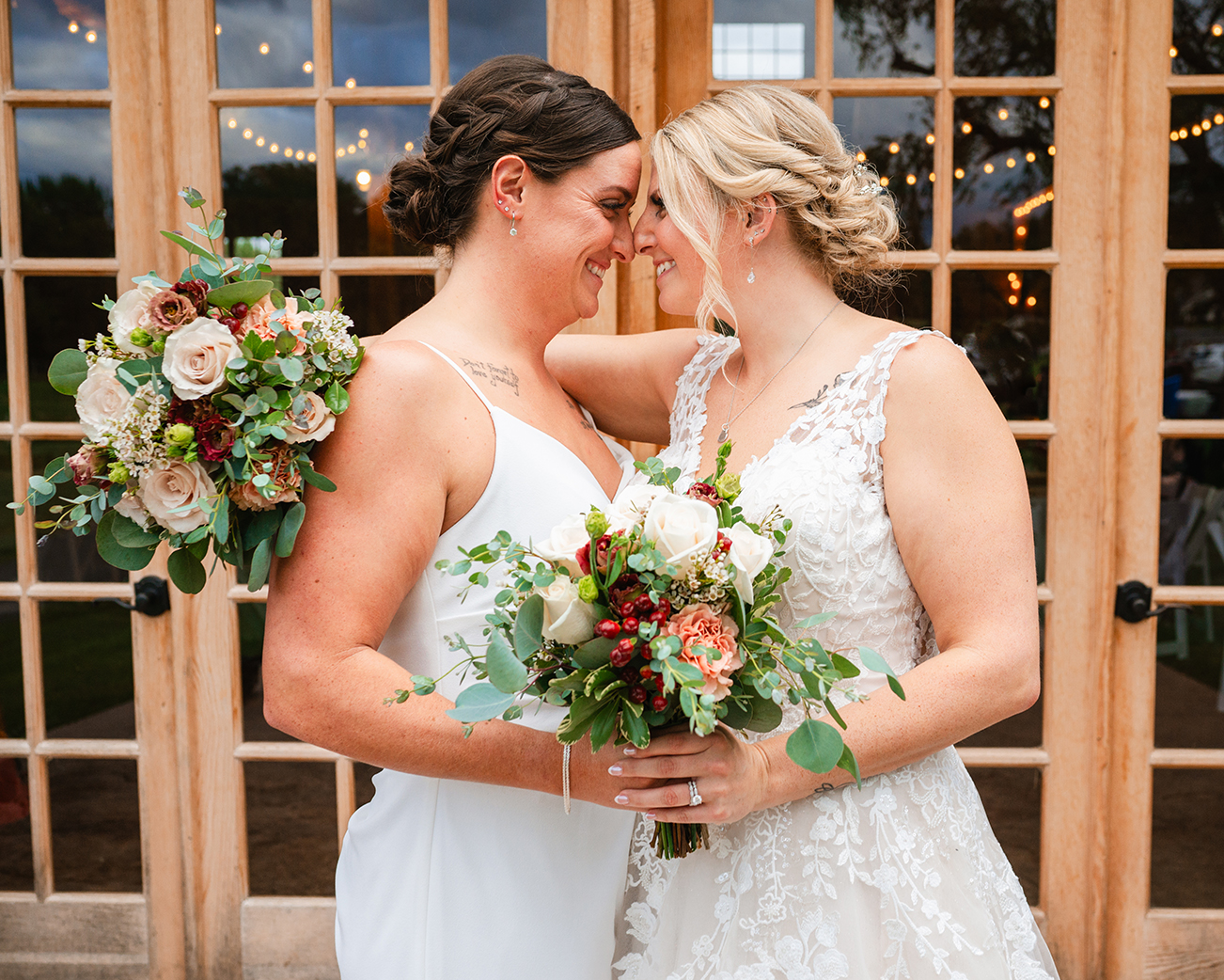 two brides in white wedding dresses with bouquets touch foreheads