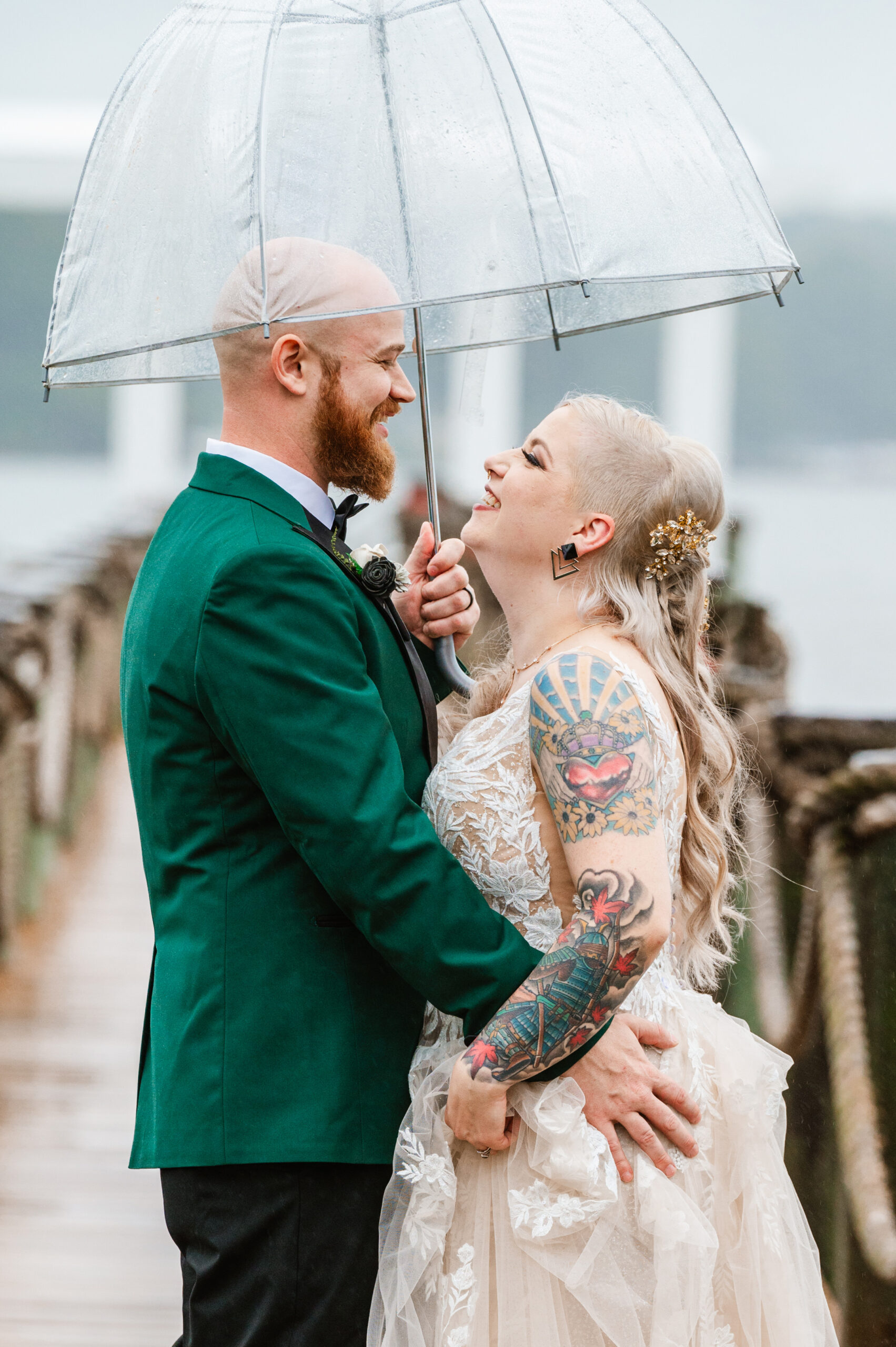 bride with colorful tattoos and groom with beard hug and smile under clear umbrella