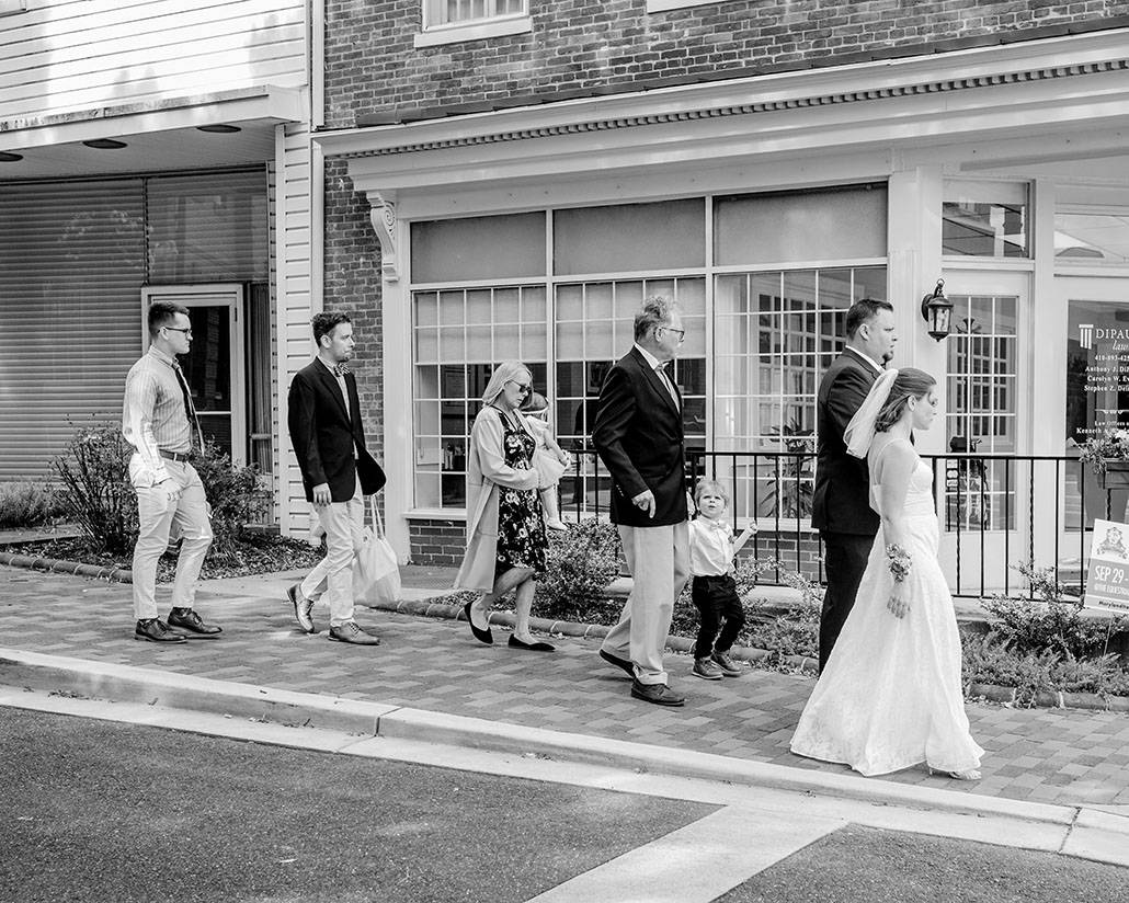 black and white image of people walking down the sidewalk with bride and groom